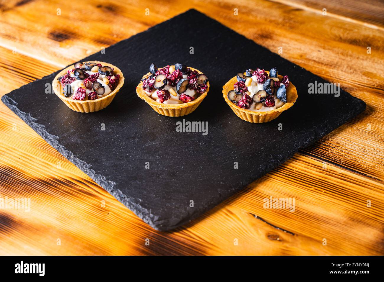 Drei Tartlets mit Hüttenkäse-Creme und Heidelbeeren auf einem schwarzen Glimmerbrett auf hölzernem Hintergrund. Hochwertige Fotos Stockfoto
