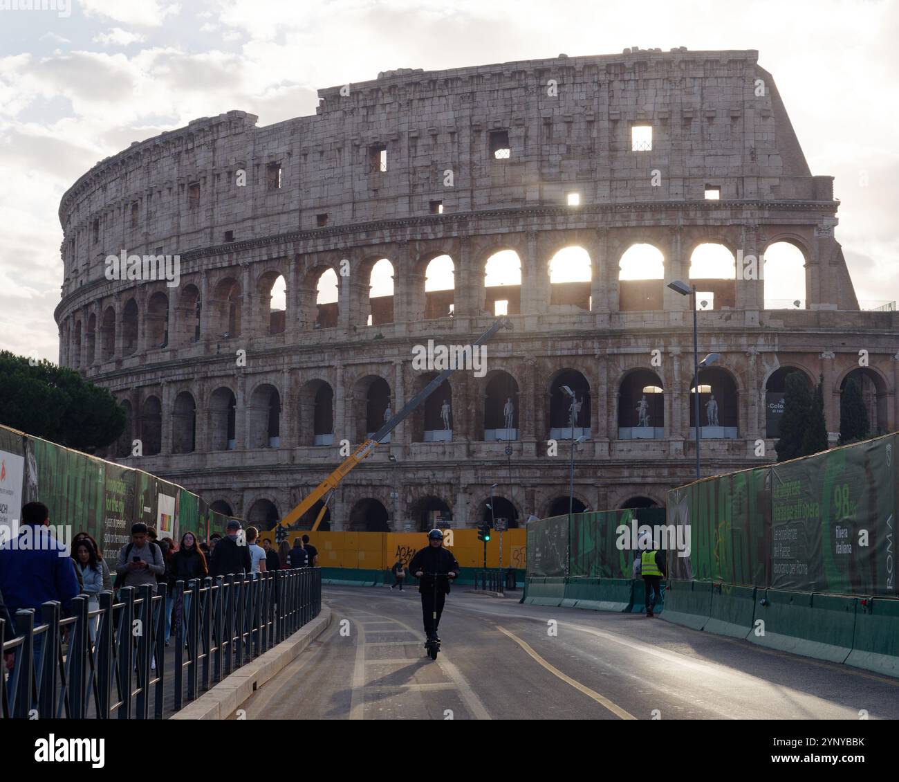 Person auf dem Roller, während die Menschen auf dem Bürgersteig vor dem Kolosseum, auch bekannt als Flavisches Amphitheater in Rom, Italien, laufen. November 2024 Stockfoto