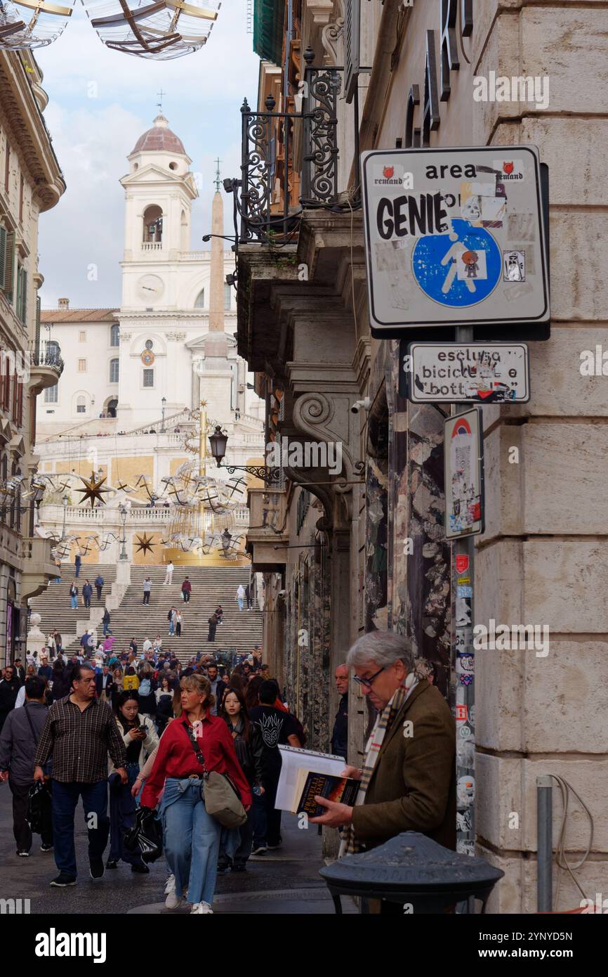 Gut gekleideter reifer Mann mit Brille liest ein Buch auf einer Straße voller Menschen mit den spanischen Stufen in Rom, Italien. November 2024 Stockfoto