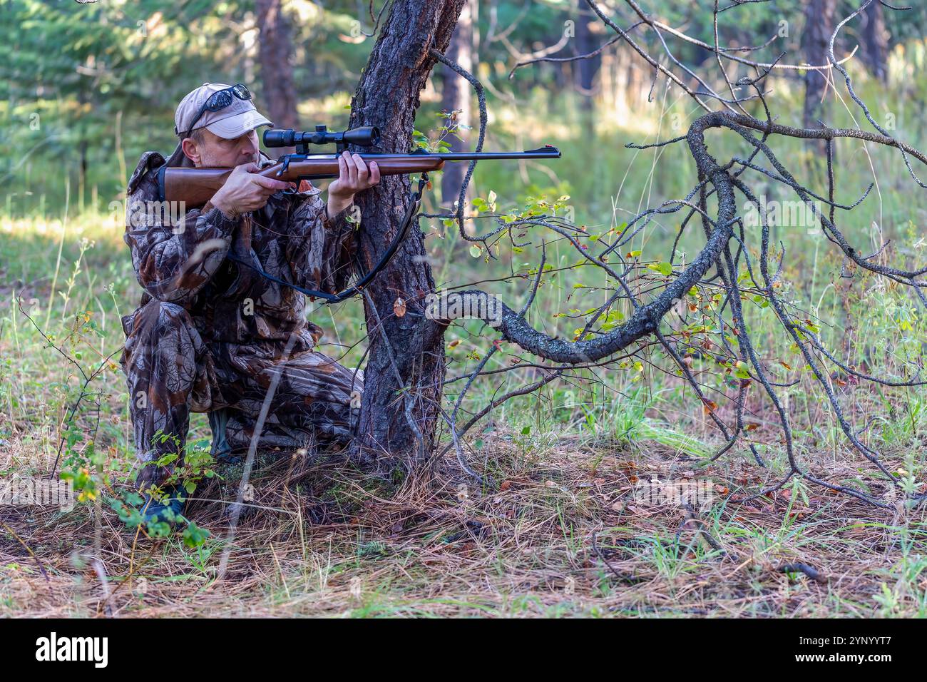 Hunter in Tarnkleidung ist in kniender Position hinter dem Baum und richtet sein Gewehr auf das Spiel, während er durch das Oszilloskop schaut. Seitenansicht. Jagen. Stockfoto