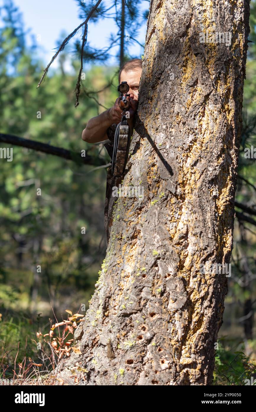 Hunter versteckt sich hinter einem großen Baum und zielt mit einem Gewehr, während er durch das Oszilloskop schaut. Bereit für die Aufnahme. Jagd im Wald. Vorderansicht... Stockfoto