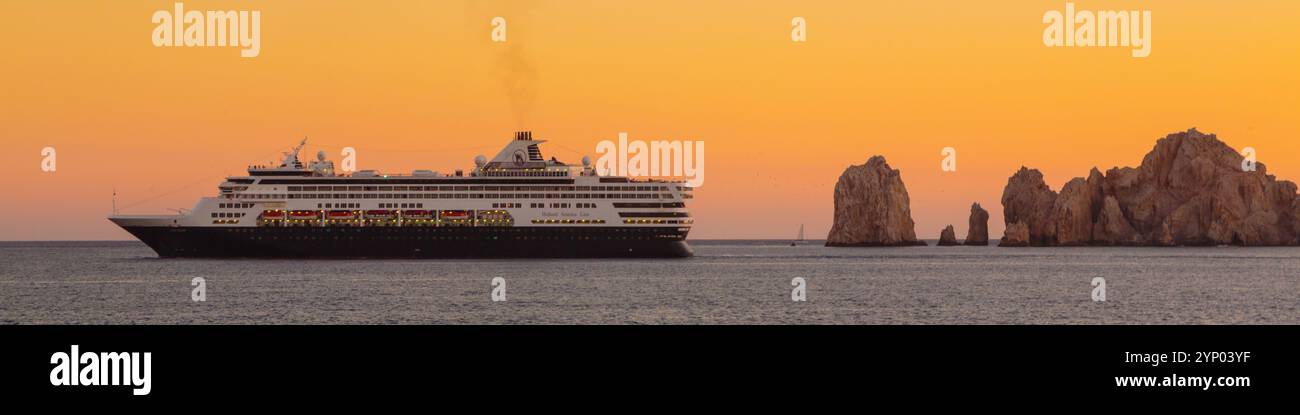 Ein großes Kreuzfahrtschiff segelt im Ozean in der Nähe eines felsigen Ufers. Der Himmel ist orange und das Wasser ist ruhig Stockfoto