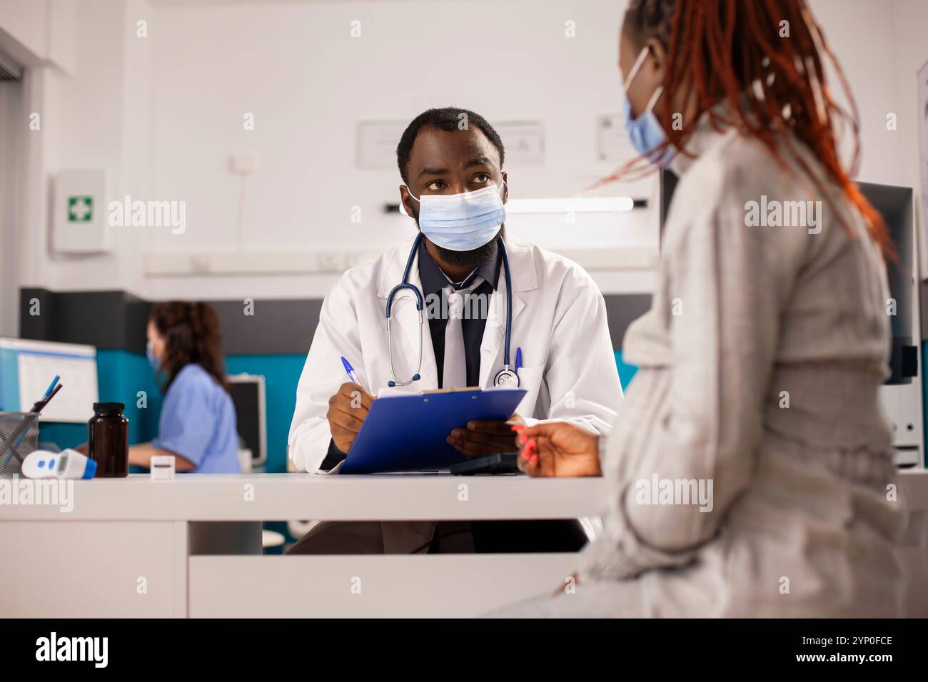 Schwangere schwarze Frau spricht mit dem Arzt am Schreibtisch, beide tragen Gesichtsmasken zum Schutz. Weibliche Krankenschwester arbeitet am Computer im Hintergrund mit Gesundheitsdokumenten, während männlicher Arzt sich um den Patienten kümmert. Stockfoto