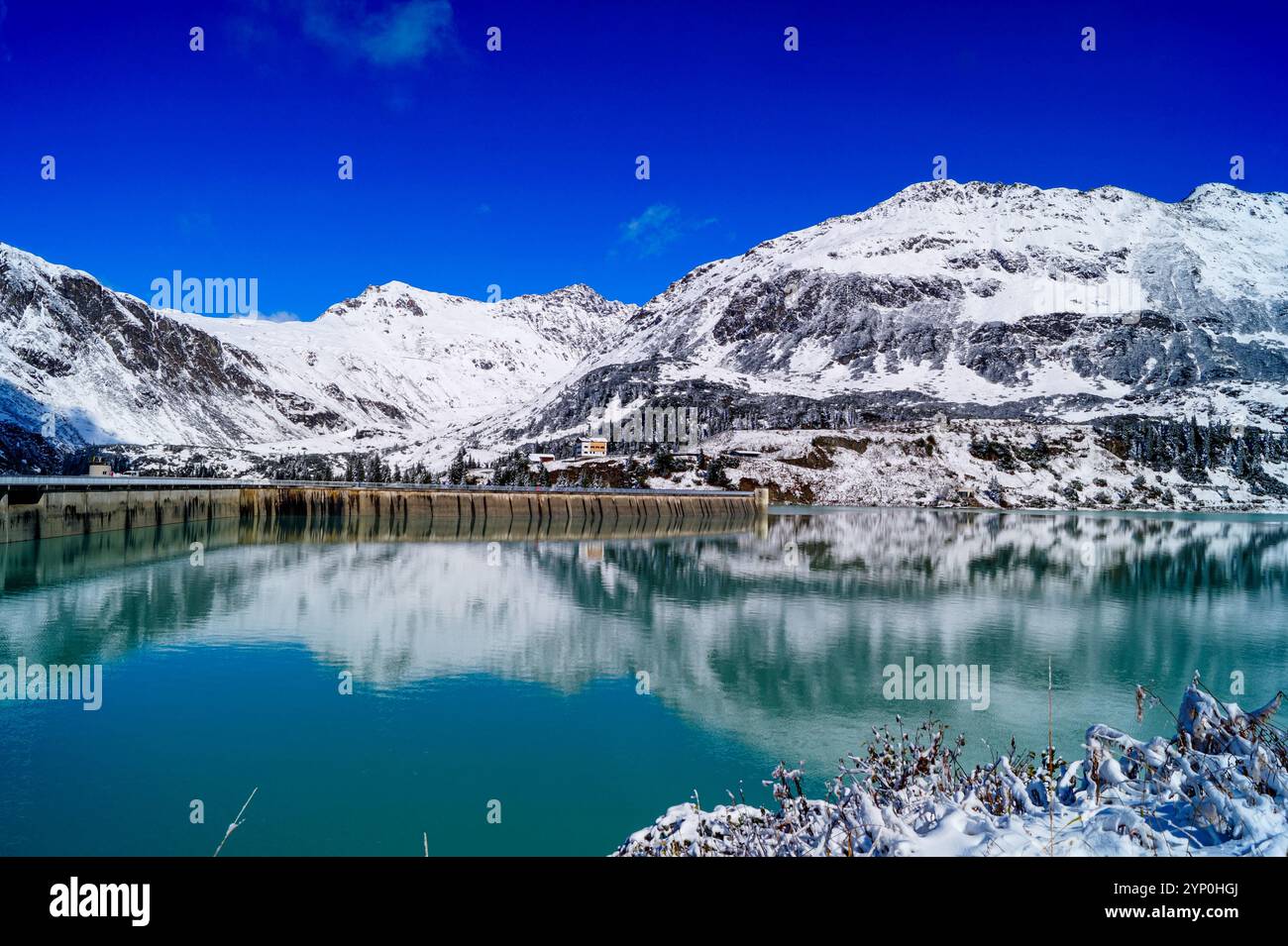 Blick auf den malerischen Alpensee mit azurblauem Wasser, Fragment eines Betondamms und Reflexionen, umgeben von schneebedeckten Bergen. Stockfoto