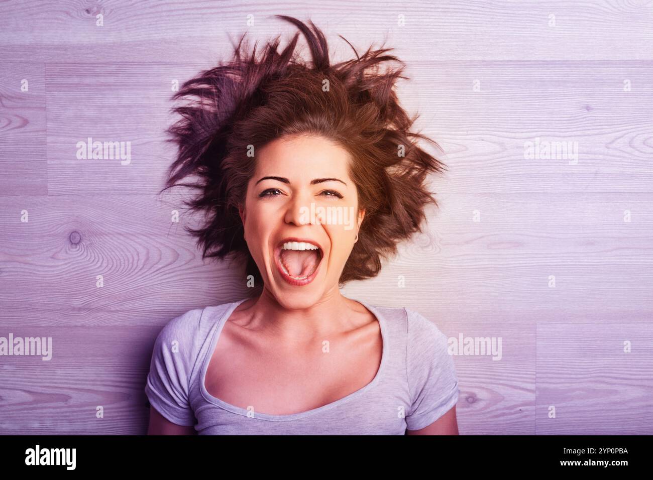 Eine junge Frau mit unordentlichem braunem Haar liegt auf einem Holzboden. Ihr Kopf ist nach hinten geneigt und der Mund weit offen, und das in einem Schrei von Frustration, Freude oder Aufregung Stockfoto