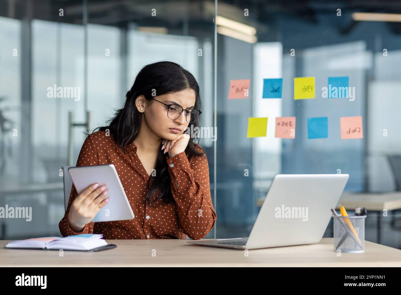 Hispanische Geschäftsfrau tief in Gedanken im modernen Büro mit Tablette. Schwerpunkt auf Notebook, umgeben von Haftnotizen, Telefon zur Hand, Darstellung der Arbeitsumgebung, Entschlossenheit, Multitasking. Stockfoto
