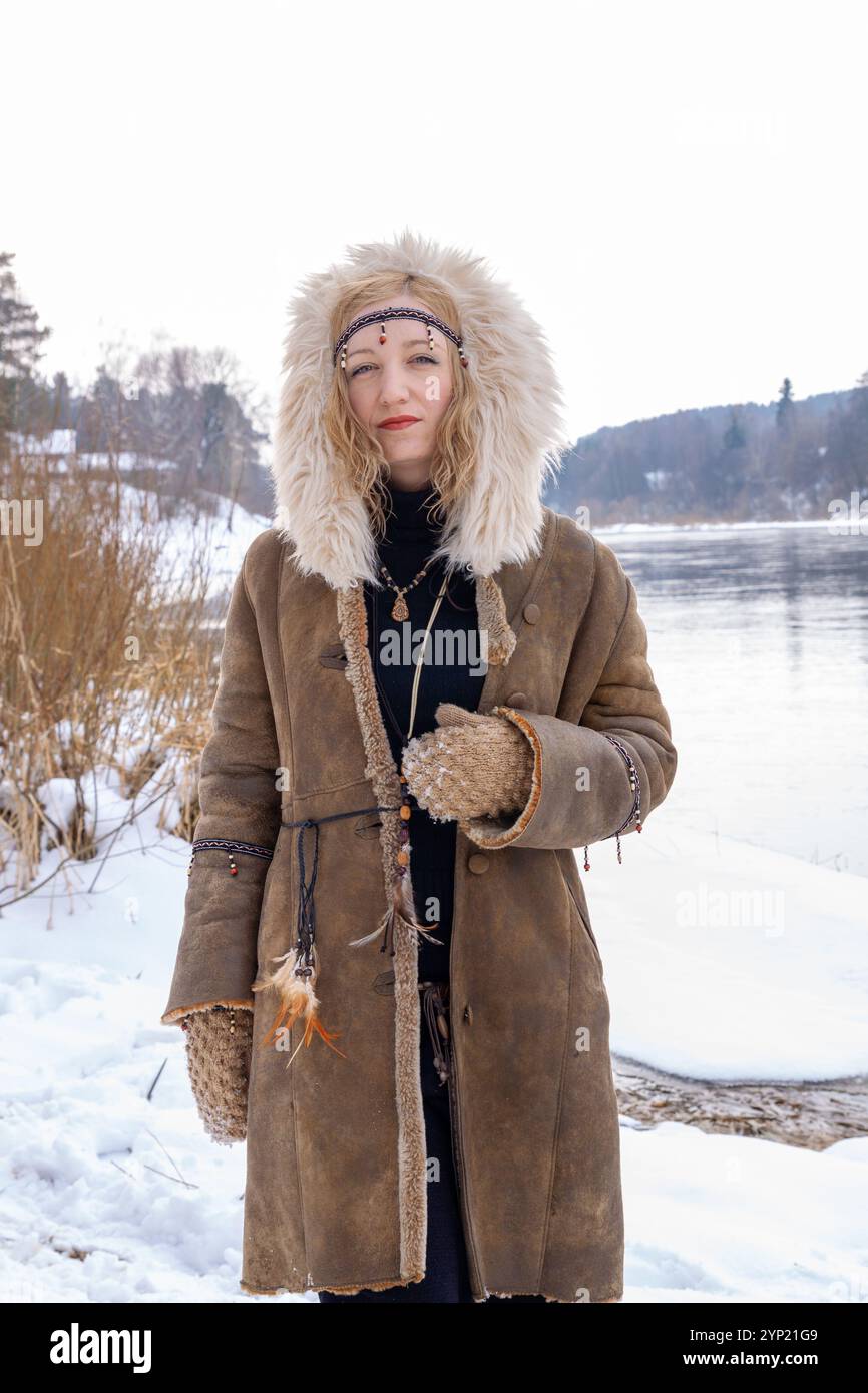 Eine junge Frau mit blonden Haaren, gekleidet in einem warmen Pelzmantel mit Accessoires im Boho-Stil, am Ufer an einem verschneiten Wintertag Stockfoto