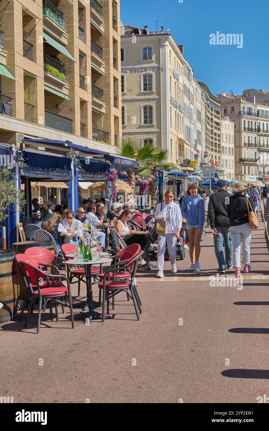 Ein Café im Freien mit Menschen an einem sonnigen Tag in Marseille. Die historischen Gebäude und die belebte Straße tragen zur charmanten und lebhaften Atmosphäre bei. Stockfoto