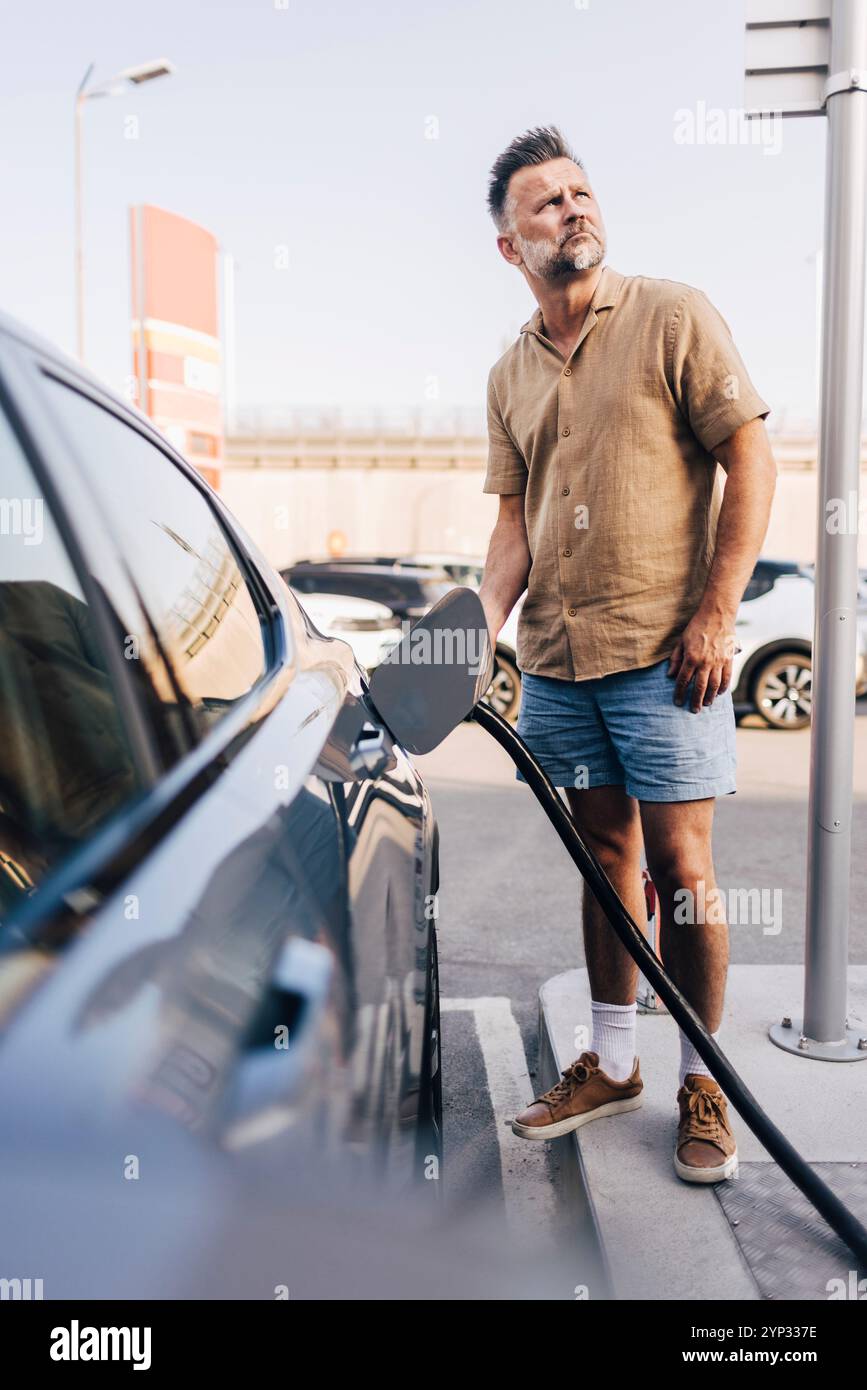 Volle Länge eines Reifen Mannes, der das Auto mit Elektrostecker an der Station auflädt Stockfoto