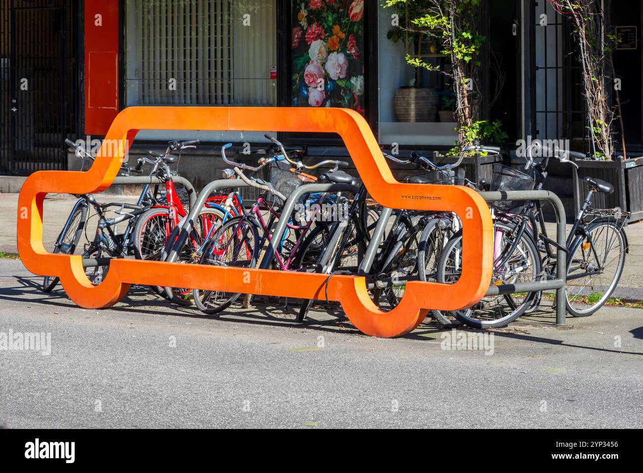 Ein Fahrradträger mit einer orangen Fassade in Form einer Autosilhouette von Cyclehoop in Malmö, Schweden, mit etwa zehn Fahrrädern, die an den Lenkern verriegelt sind. Stockfoto