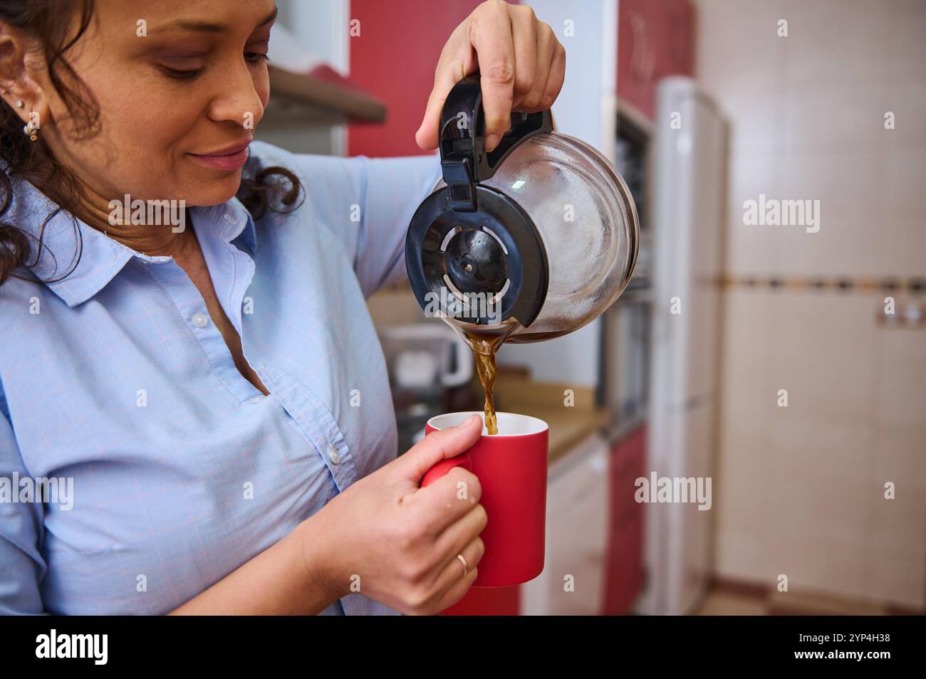 Eine Frau in einem hellblauen Hemd gießt mit einem friedlichen Lächeln dampfenden Kaffee in eine rote Tasse. Die warme Küchenkulisse sorgt für ein heimeliges Ambiente und fängt einen Mo ein Stockfoto