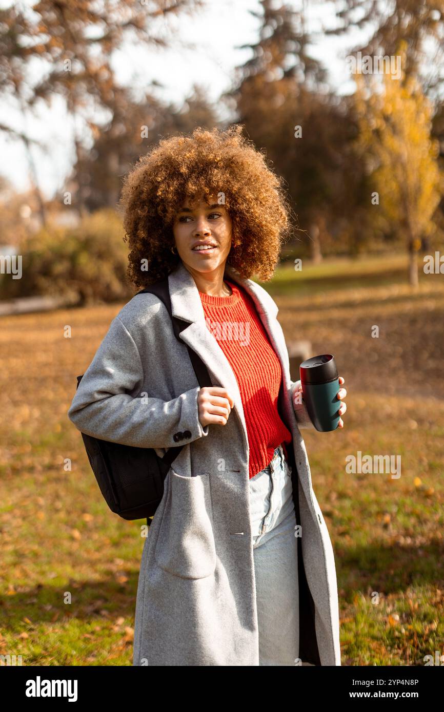 Eine fröhliche Frau mit lockigem Haar schlendert durch einen sonnigen Park, der mit bunten Herbstblättern geschmückt ist, hält eine warme Tasse Kaffee in der Hand und trägt eine stilvolle Stockfoto