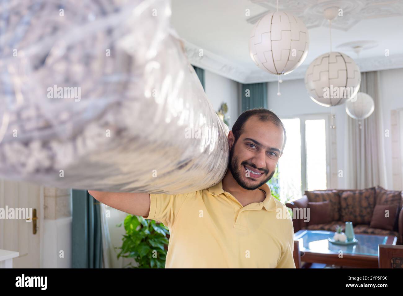 Fröhlicher Mann in einem gelben Hemd, der einen gerollten Teppich über der Schulter trägt, in einem stilvollen Wohnzimmer, umgeben von elegantem Dekor. Erfasst eine Mo Stockfoto