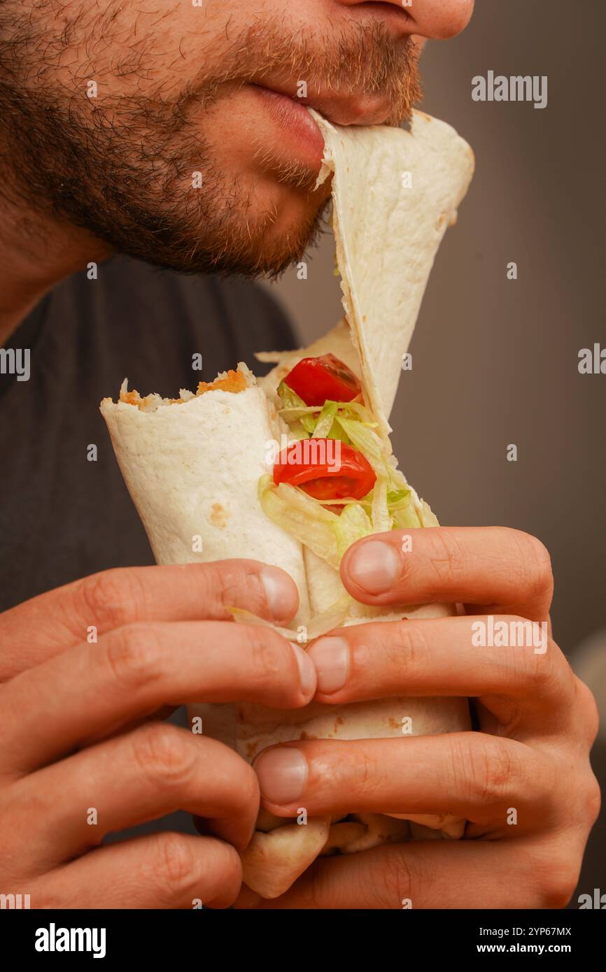 Ein Mann mit einem kleinen Bart beißt in ein Pita-Brötchen, gefüllt mit Fleisch, frischem Gemüse und Salat. Das Foto, das in der Mitte der Bewegung aufgenommen wurde, betont das Deliciou Stockfoto