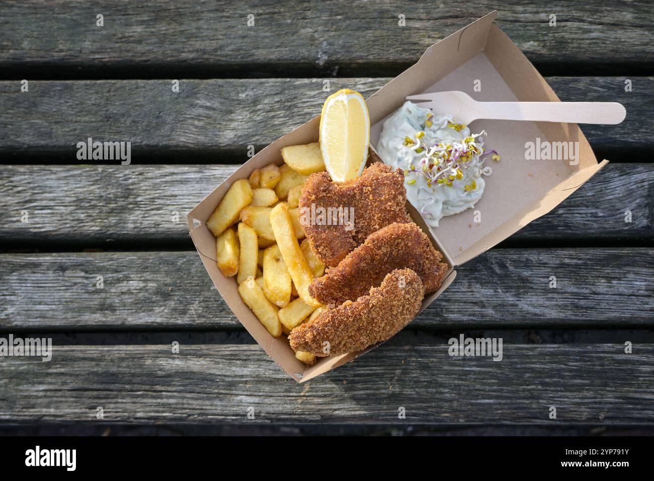 Fish and Chips mit Zitronenscheibe, Tatarsauce und Sprossen in einer Schachtel aus Pappe auf einem verwitterten Holzdock, Fisch und Meeresfrüchte-Snack an der Küste, Kopierraum, Stockfoto