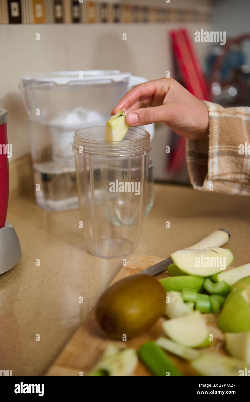 Eine Person bereitet einen grünen Smoothie zu, indem sie Apfelscheiben und Kiwi in einen Mixbecher gibt. Frische Zutaten wie Sellerie und Apfel ruhen auf einem Schneideschwein Stockfoto