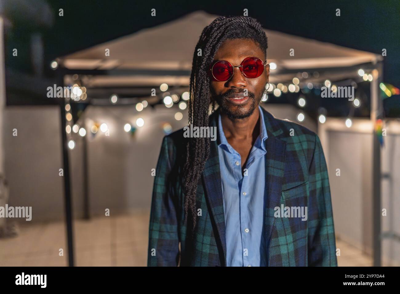 Porträt eines coolen afrikanischen Mannes mit Afro-Haarsträhnen und farbenfroher Vintage-Jacke und Sonnenbrille, der bei einer Nachtparty auf einer Terrasse steht Stockfoto