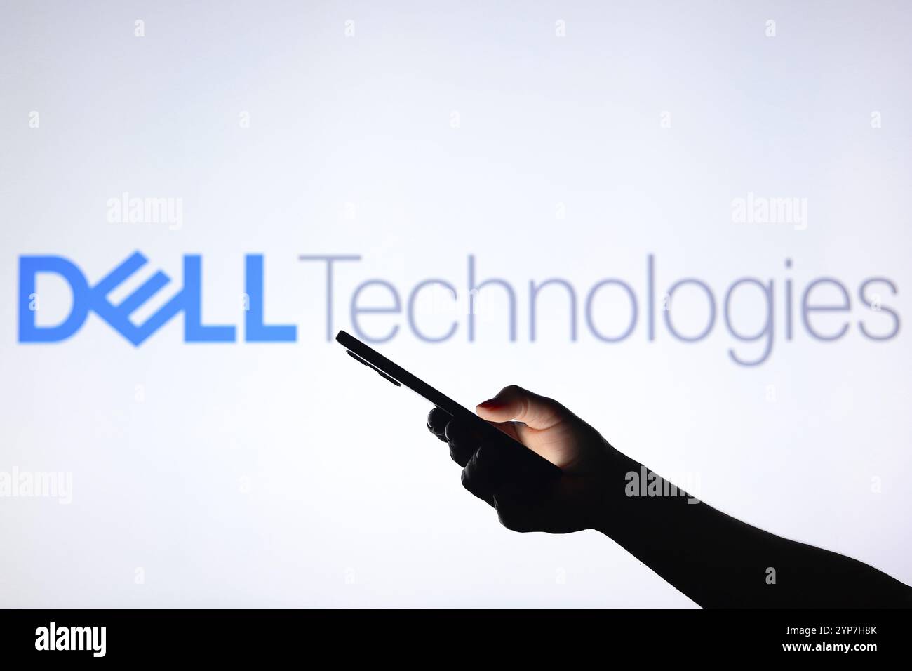 In dieser Fotoabbildung ist das Dell Technologies Logo im Hintergrund mit einer Silhouettenhand zu sehen, die ein Smartphone hält Stockfoto