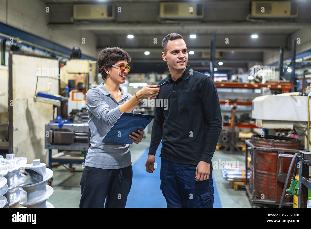 Weibliche Managerin der Fabrik spricht und erteilt Anweisungen an männliche Arbeiter, während sie das Klemmbrett hält Stockfoto