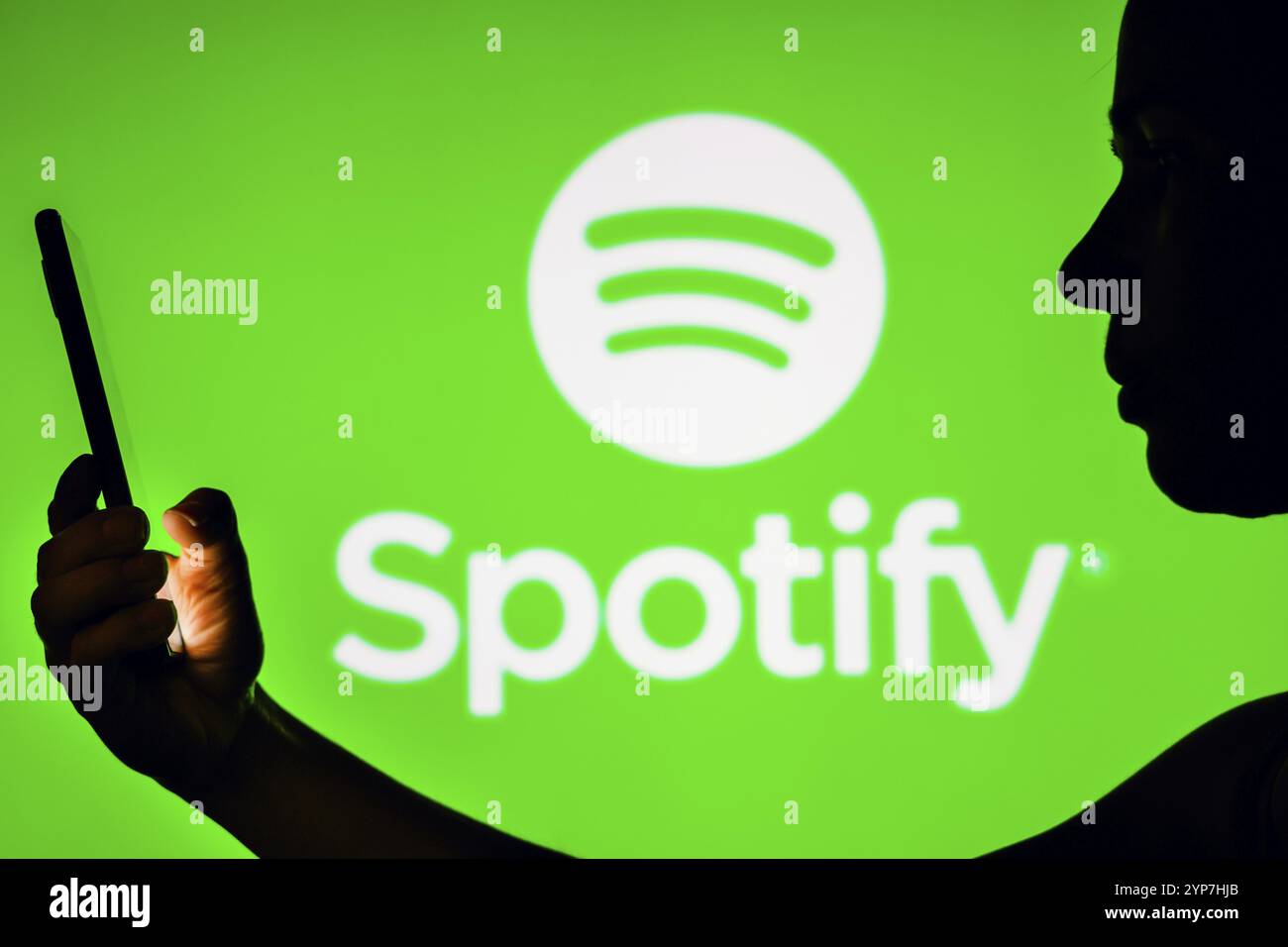 In dieser Fotoabbildung hält die Silhouette einer Frau ein Smartphone mit dem Spotify-Logo im Hintergrund Stockfoto