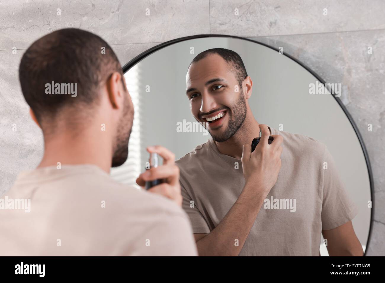 Lächelnder Mann, der Parfüm neben dem Spiegel im Badezimmer sprüht Stockfoto