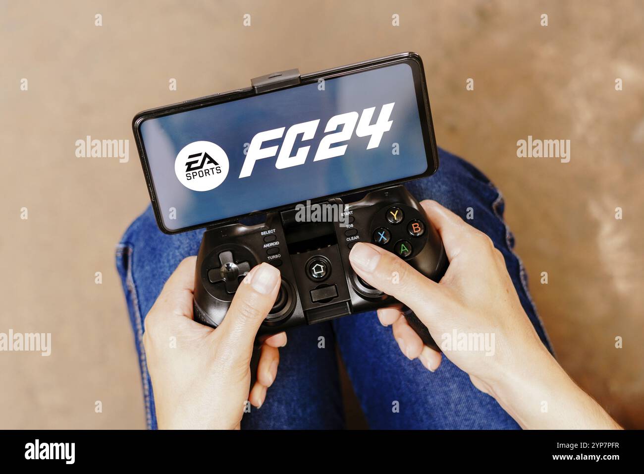 In dieser Fotoabbildung wird eine Person, die auf einem Joystick spielt, und das EA Sports FC 24-Logo auf einem Smartphone-Bildschirm angezeigt Stockfoto