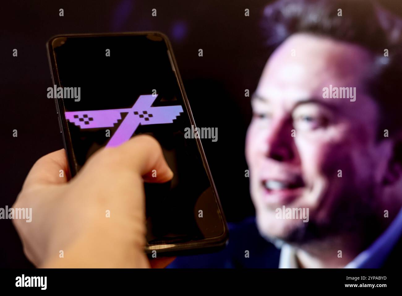 In dieser Abbildung ist das Grok-Münzlogo auf einem Smartphone-Bildschirm vor einem Computerbildschirm zu sehen, der ein Porträt von Elon Musk zeigt. Stockfoto