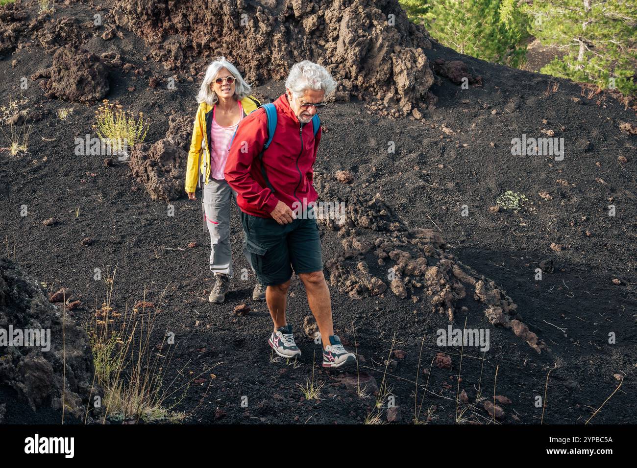 Aktives Seniorenpaar mit farbenfrohen Jacken, das vulkanisches Gelände erkundet. Natürlicher ehrlicher Moment abenteuerlicher Rentner, die auf einem dramatischen Lavafeld wandern, EM Stockfoto