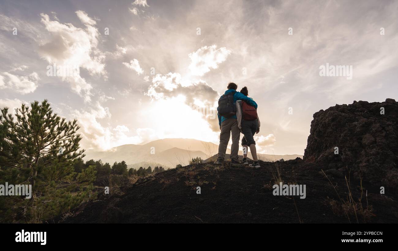 Paare stehen zusammen auf vulkanischer Felsformation und bewundern die dramatische Berglandschaft bei Sonnenuntergang. Silhouettenfiguren vor goldenen Himmelseinfangern Stockfoto
