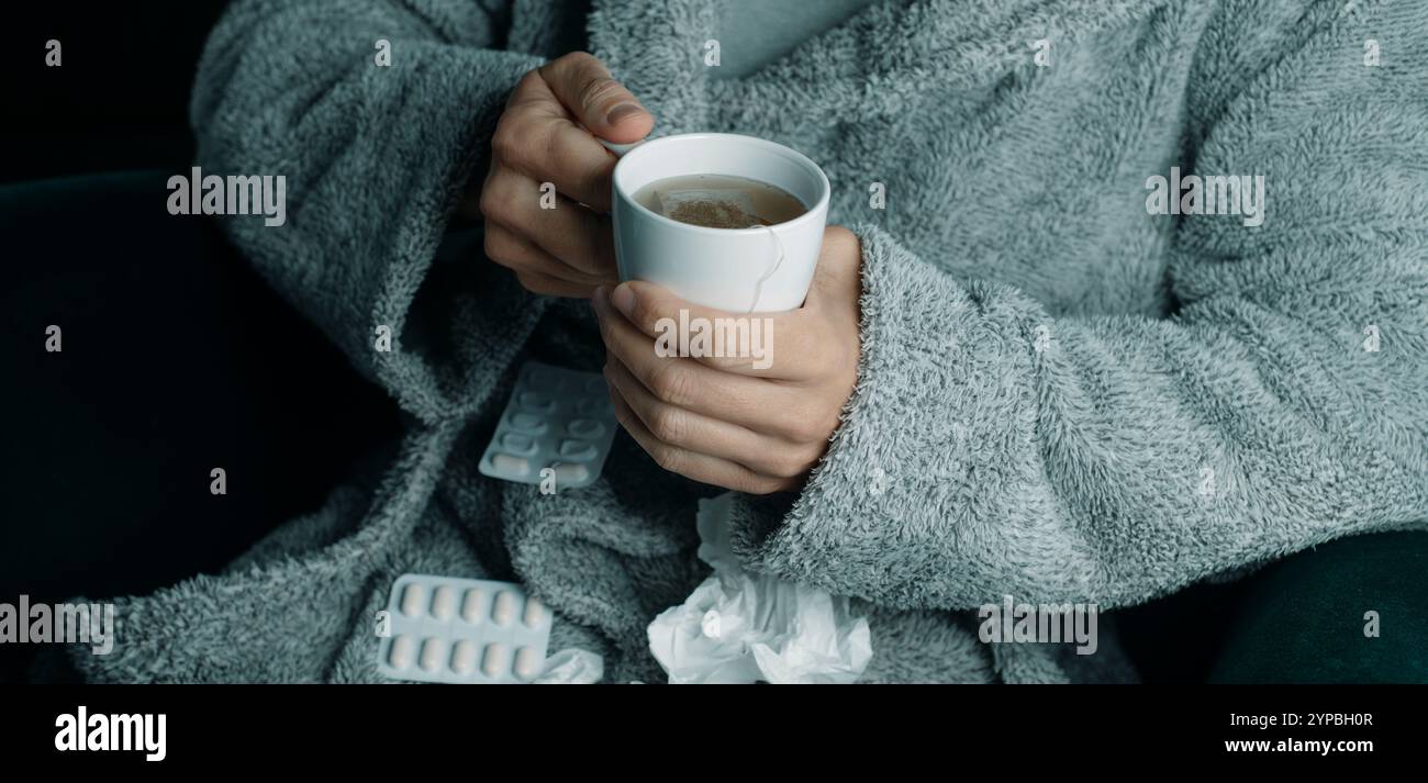 Detail eines kranken Mannes, der sich mit einer Tasse heißem Kräutertee aufwärmt, in einen flauschigen grauen Hausmantel gewickelt, zu Hause Stockfoto