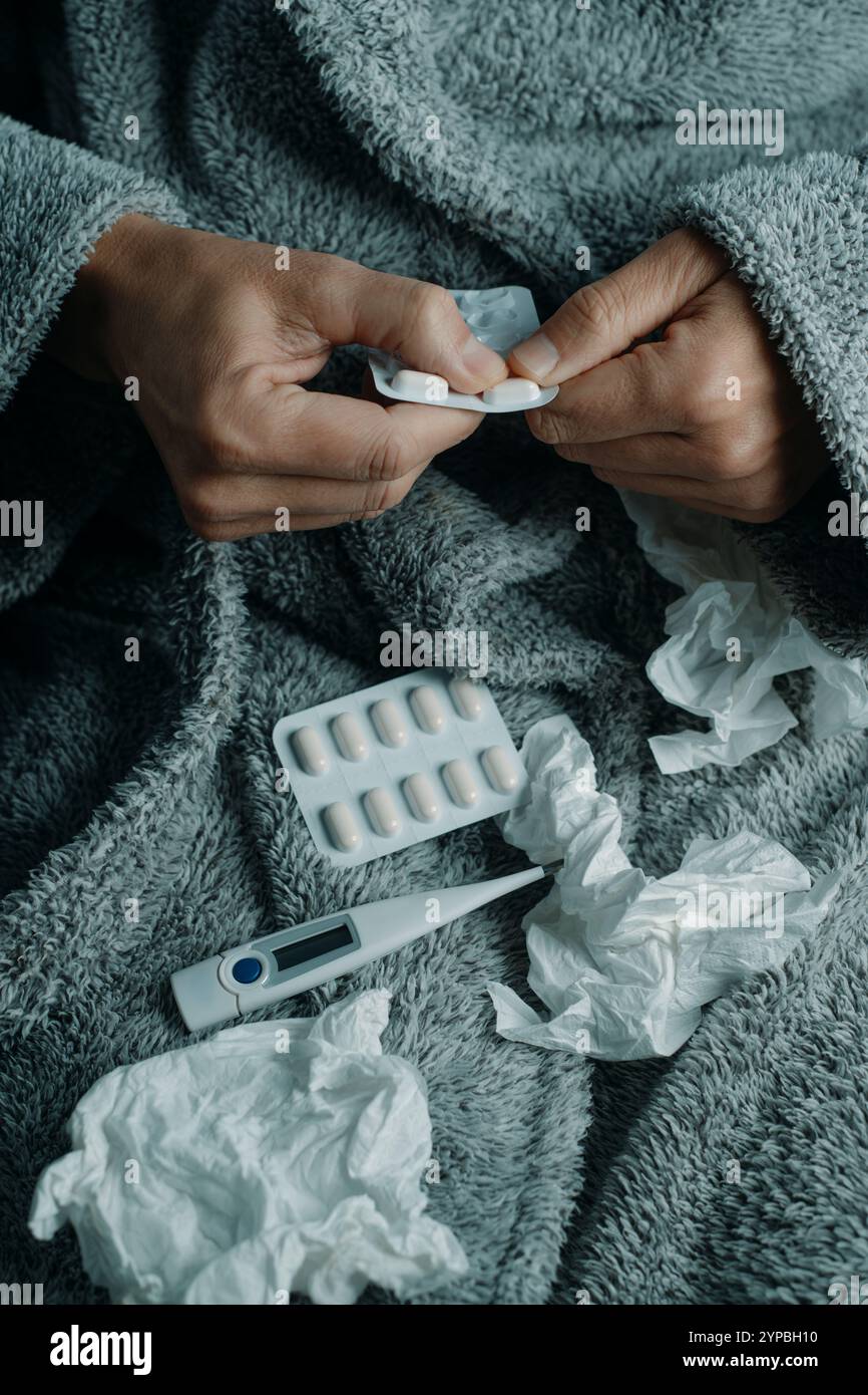 Ein kranker Mann zu Hause, gewickelt in einen flauschigen grauen Hausmantel, ist dabei, eine Pille zu nehmen, umgeben von gebrauchten Taschentüchern Stockfoto