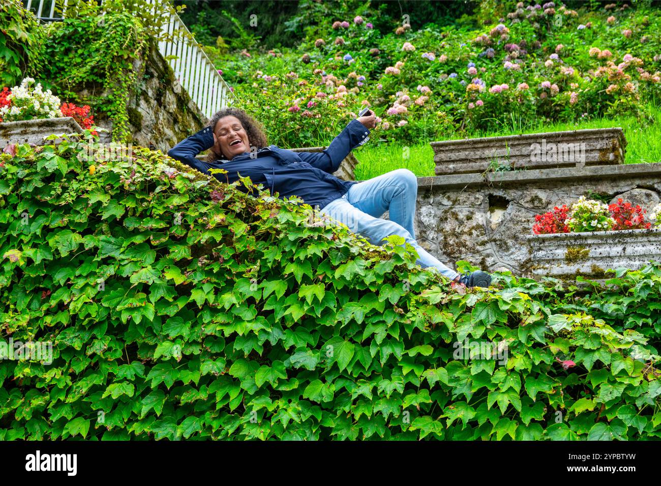 Amüsante, schöne südamerikanische Frau, mittleres Alter mit lockigen Haaren, zwischen Blättern und Blumen gelegen. Stockfoto