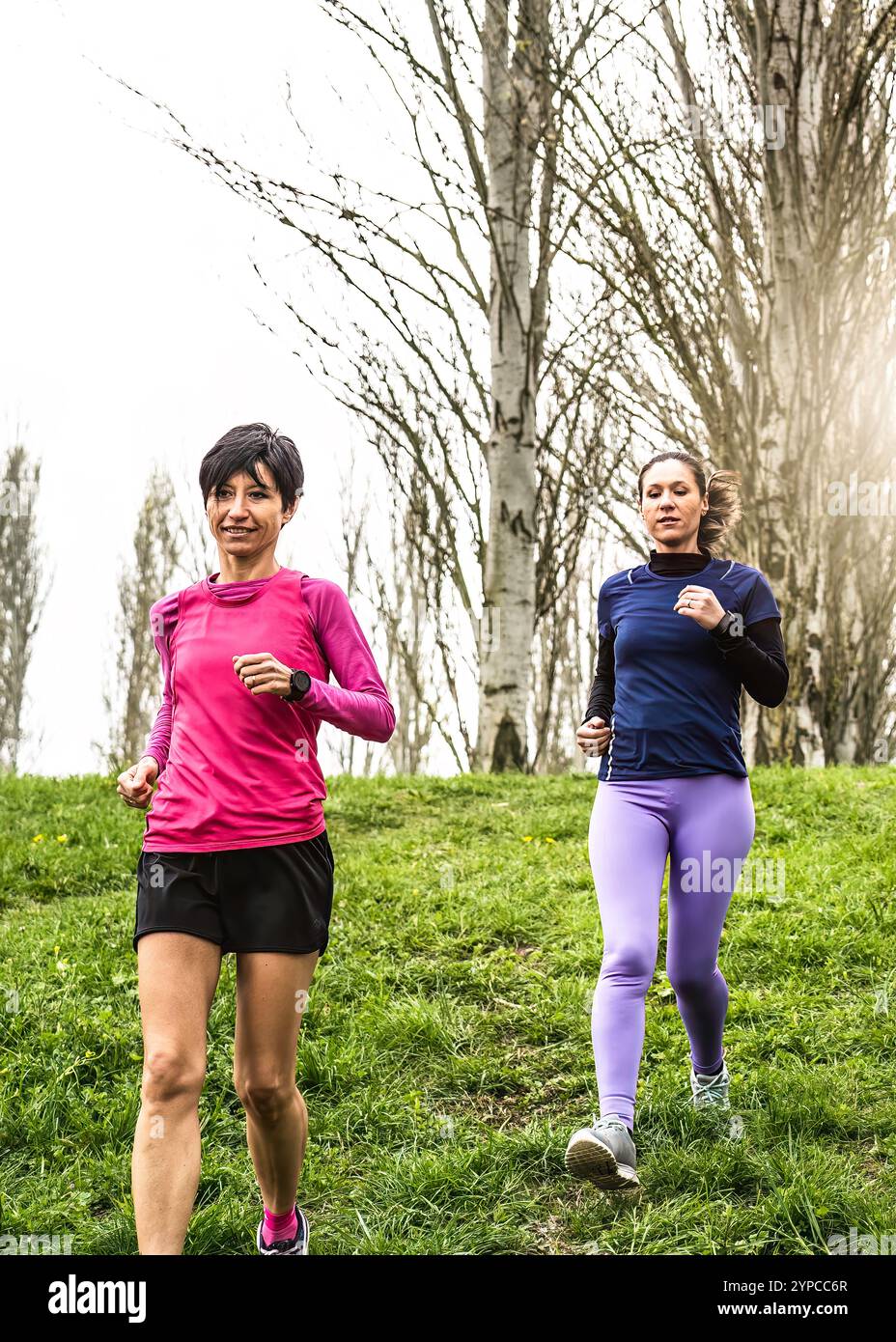 Zwei Frauen joggen auf einem grasbewachsenen Parkweg, umgeben von Bäumen. Konzentrieren Sie sich auf eine Outdoor-Fitness-Aktivität im Frühling, um Gesundheit, Bewegung und zu fördern Stockfoto