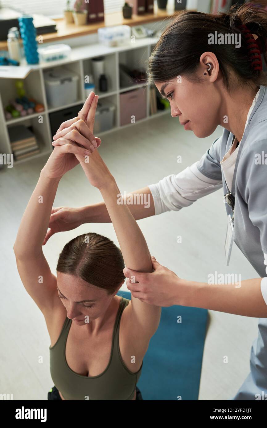 Medizinisches Fachpersonal, das Patienten mit Dehnungsübungen in einer gut beleuchteten, organisierten Klinik unterstützt. Regale mit verschiedenen medizinischen Vorräten und Büchern im Hintergrund sichtbar Stockfoto