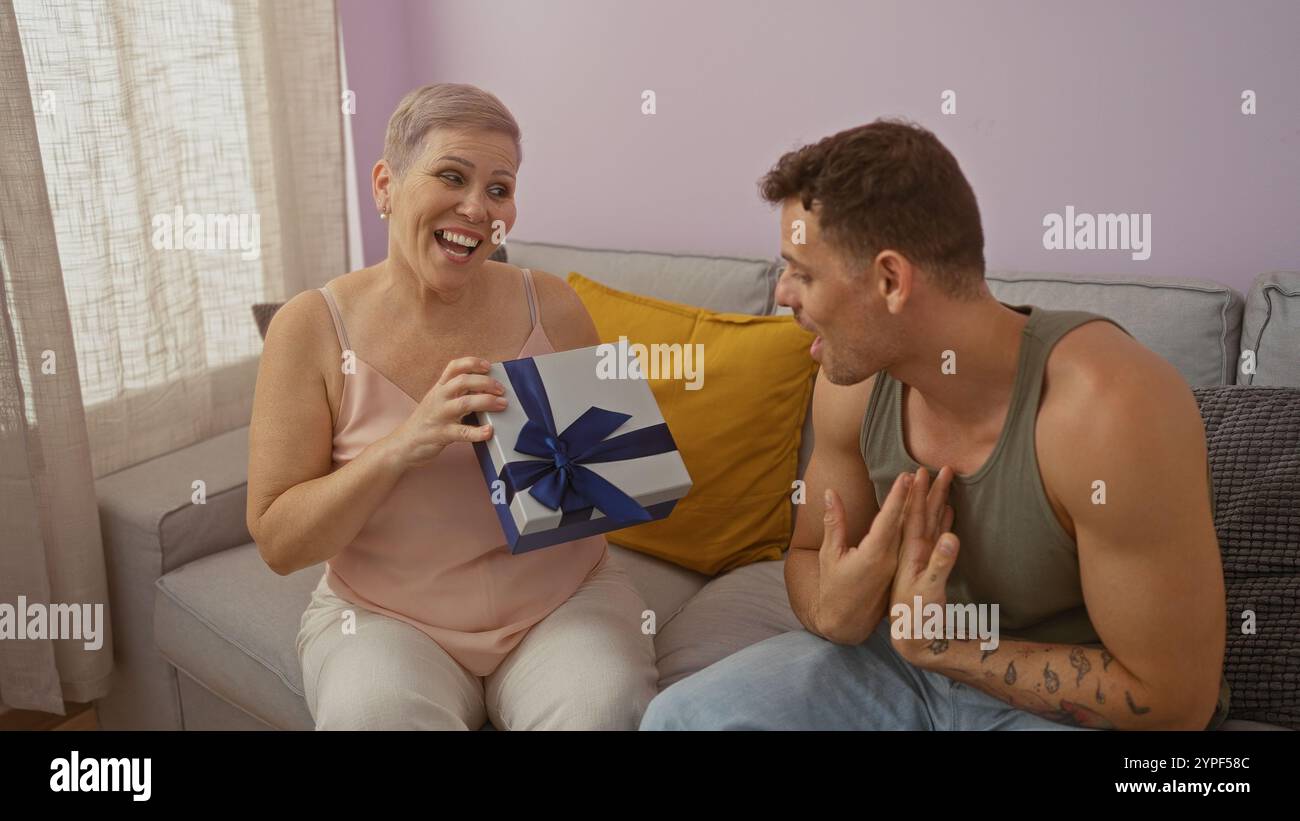 Die Mutter empfängt glücklich ein Geschenk von ihrem Sohn im Wohnzimmer mit einem großen Lächeln, zeigt ihre starke Familienbindung und Liebe, sitzt zusammen auf einem Sof Stockfoto