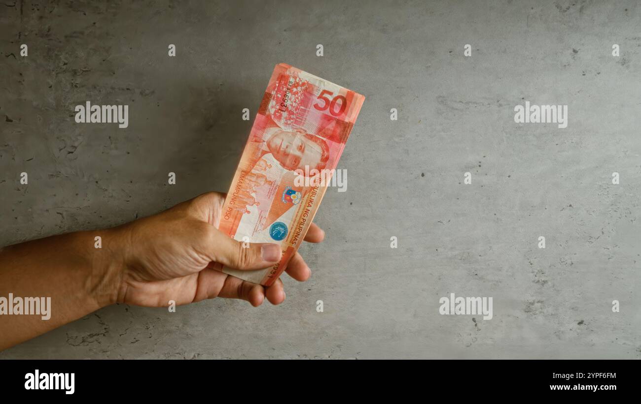 Ein Mann, der eine philippinische Banknote mit 50 Peso vor einem texturierten Betonhintergrund hält und die Währung in der Hand mit lebendigen Details des Banknotendesigns zeigt Stockfoto