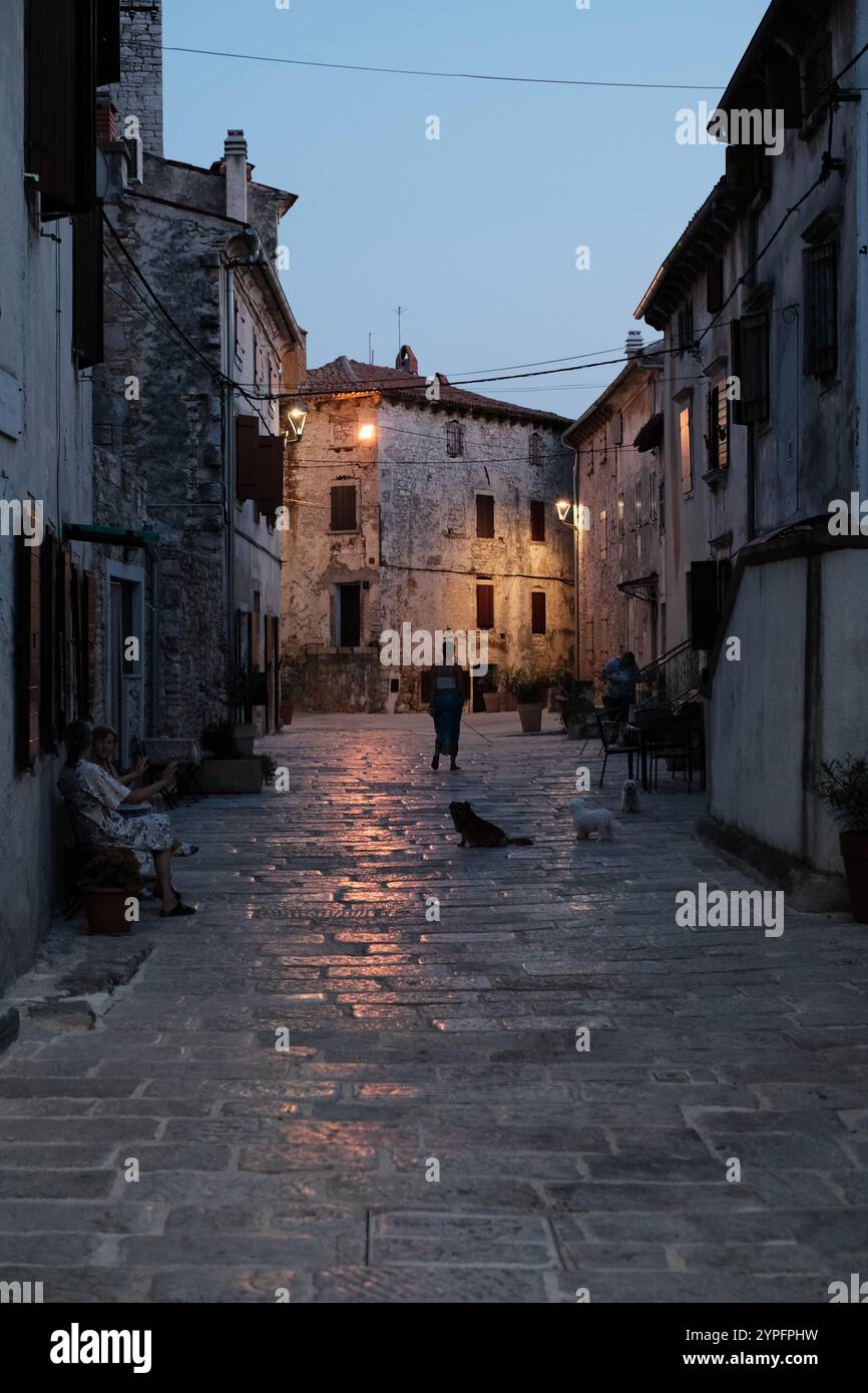 Altstadtstraße in Bale, Istrien am Abend. Leute, die vor alten Steinhäusern sitzen. Straßenlaternen reflektieren die gepflasterte Straße. Stockfoto