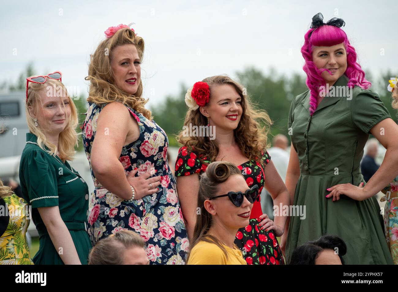 Frauengruppen präsentieren bei einem lebhaften Treffen im Freien lebendige Retro-Outfits. Ihr selbstbewusstes Lächeln und ihre verspielten Posen unterstreichen den Geist des Abends Stockfoto