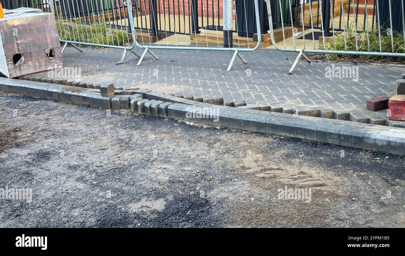 Ein Abschnitt des neu verlegten Gehwegs weist beschädigte Bordsteinkanten und Pflastersteine auf. Lose Steine und Schmutz sind in der Nähe verstreut. Temporäre Metallbarrieren sind VI Stockfoto