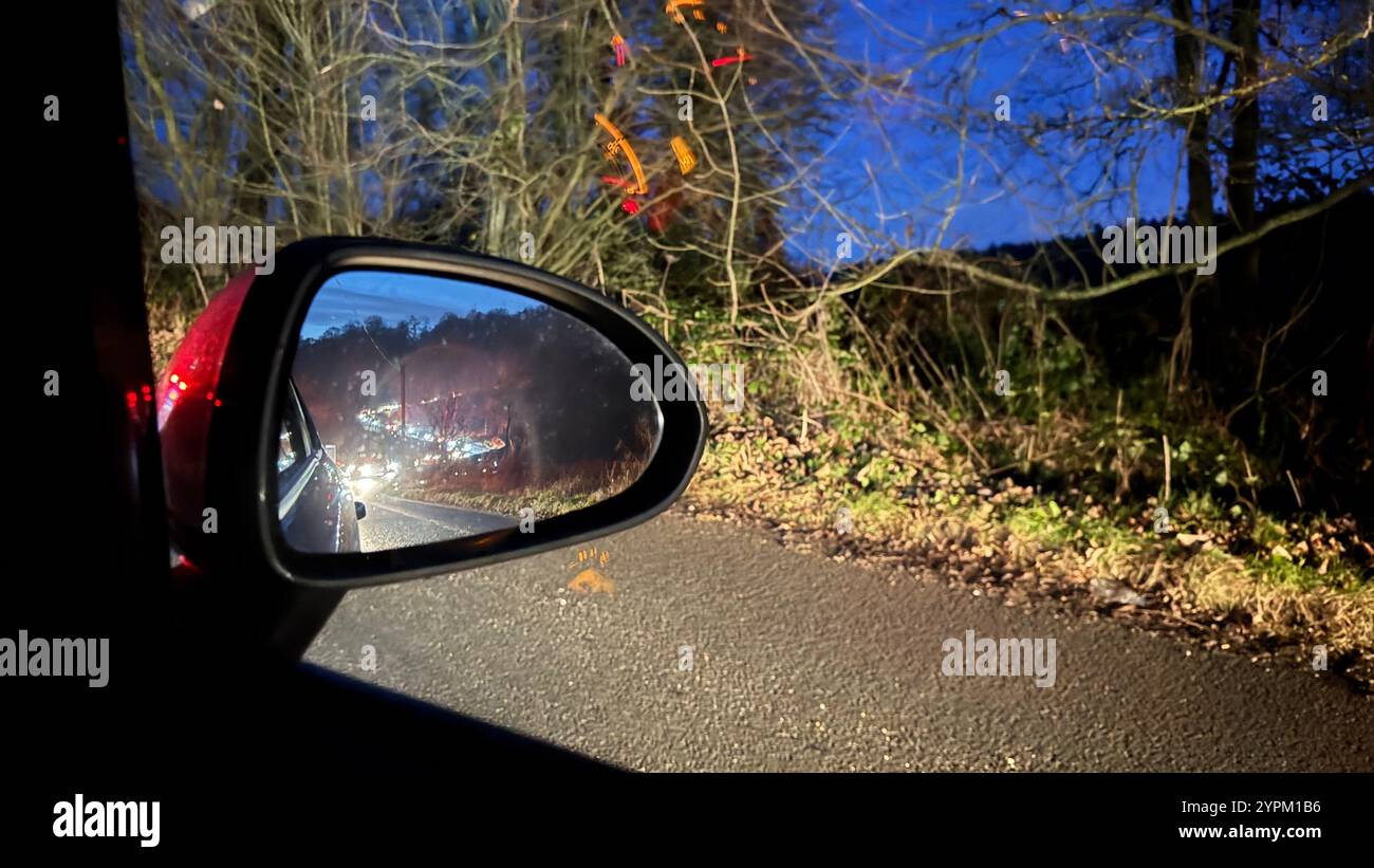 Der Seitenspiegel eines Autos reflektiert eine nächtliche Straßenszene mit verschwommenem Verkehr und Bäumen. Die Straße ist dunkel, der Himmel dämmerblau. Vegetation säumt den Ro Stockfoto