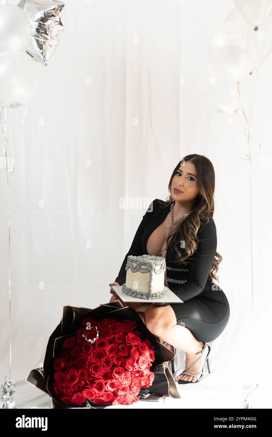 Eine Frau posiert mit einem Kuchen und einem Strauß roter Rosen. Der Kuchen wird vor ihr auf einen Tisch gestellt, und die Rosen sind in einer Vase. Das Konzept des Celeb Stockfoto