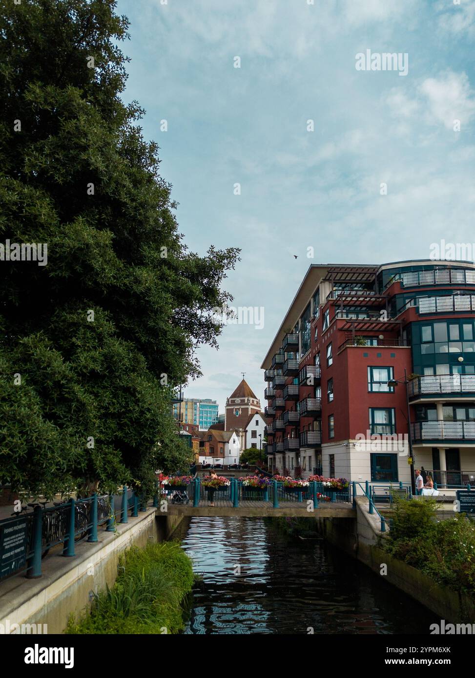 Bezaubernder Blick auf den Fluss mit modernen Apartments, üppigem Grün und einer historischen Kirche in einer ruhigen Stadtlandschaft Stockfoto
