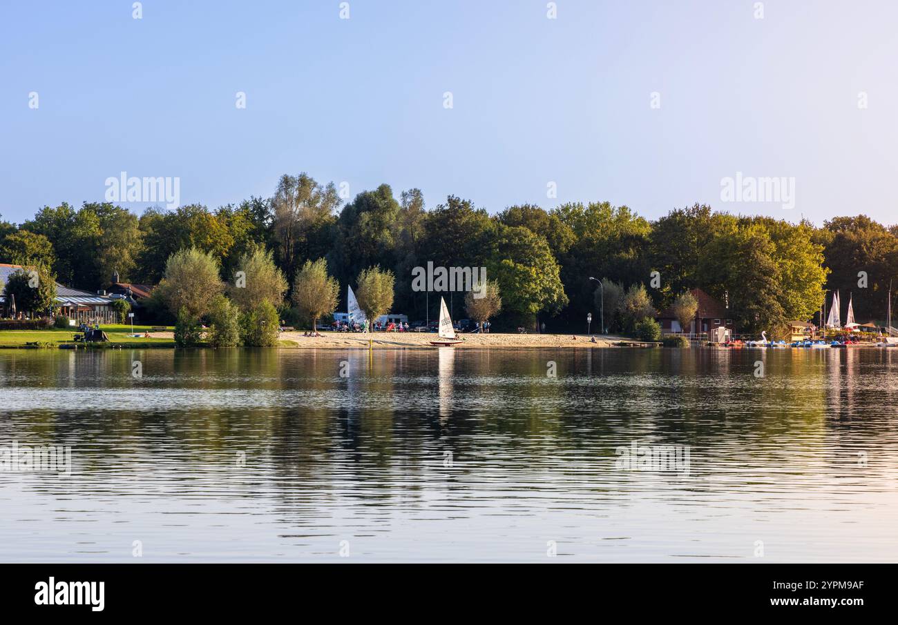 Ein ruhiger Blick auf den See mit einem Sandstrand, Segelbooten und üppigen grünen Bäumen unter dem Himmel, die eine Outdoor-Szene zeigen. Gronau, Deutschland, 2024 Stockfoto