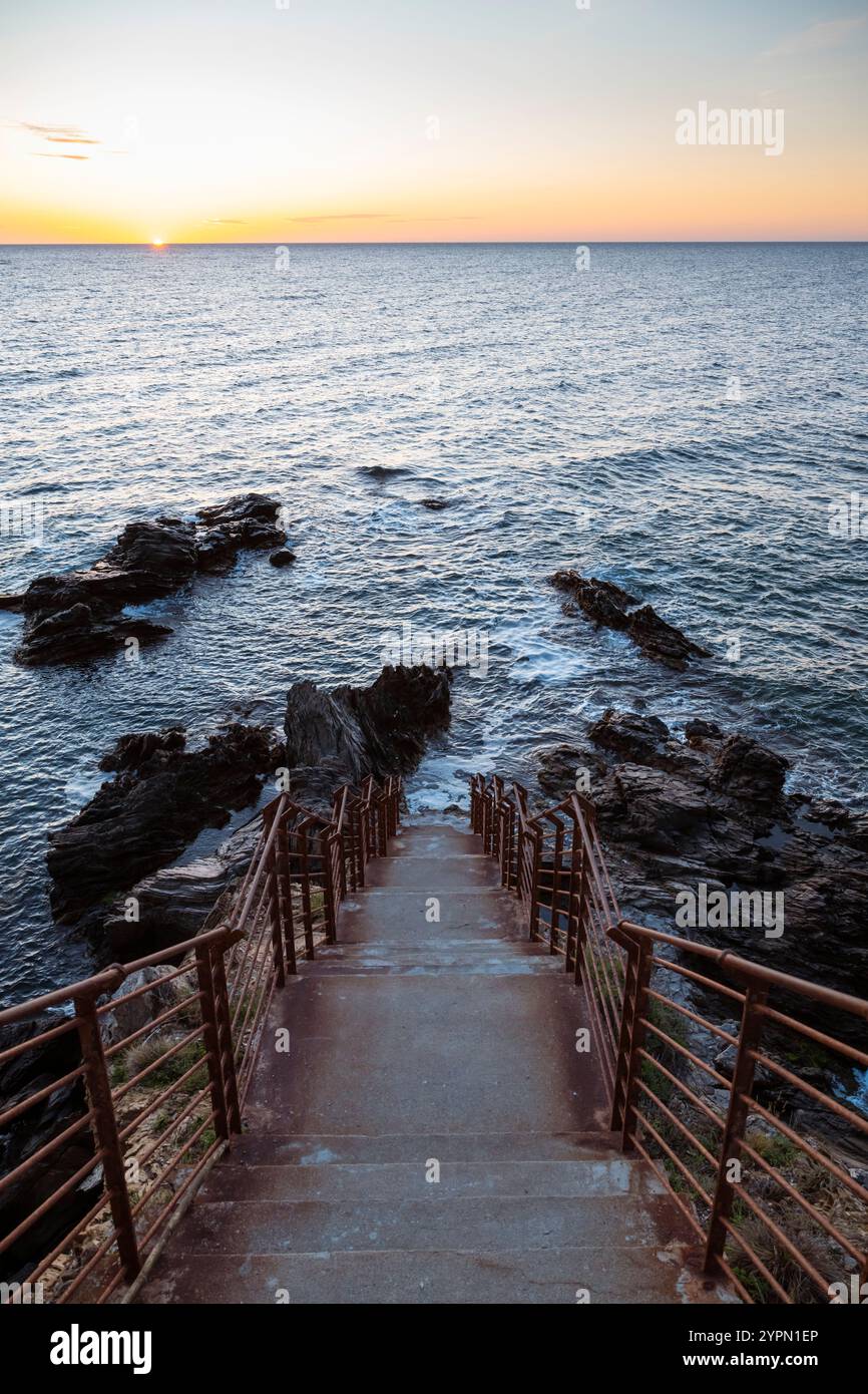 Sonnenaufgang über einer Bucht mit zerklüfteten Felsen, Betontreppe und rostigem Geländer an der Mittelmeerküste in der Nähe von Collioure, Cote Vermeille, Okzitanien Stockfoto