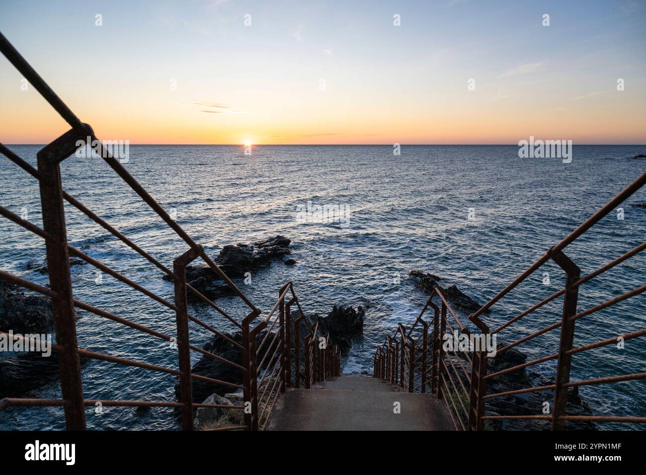 Sonnenaufgang über einer Bucht mit zerklüfteten Felsen, Betontreppe und rostigem Geländer an der Mittelmeerküste in der Nähe von Collioure, Cote Vermeille, Okzitanien Stockfoto