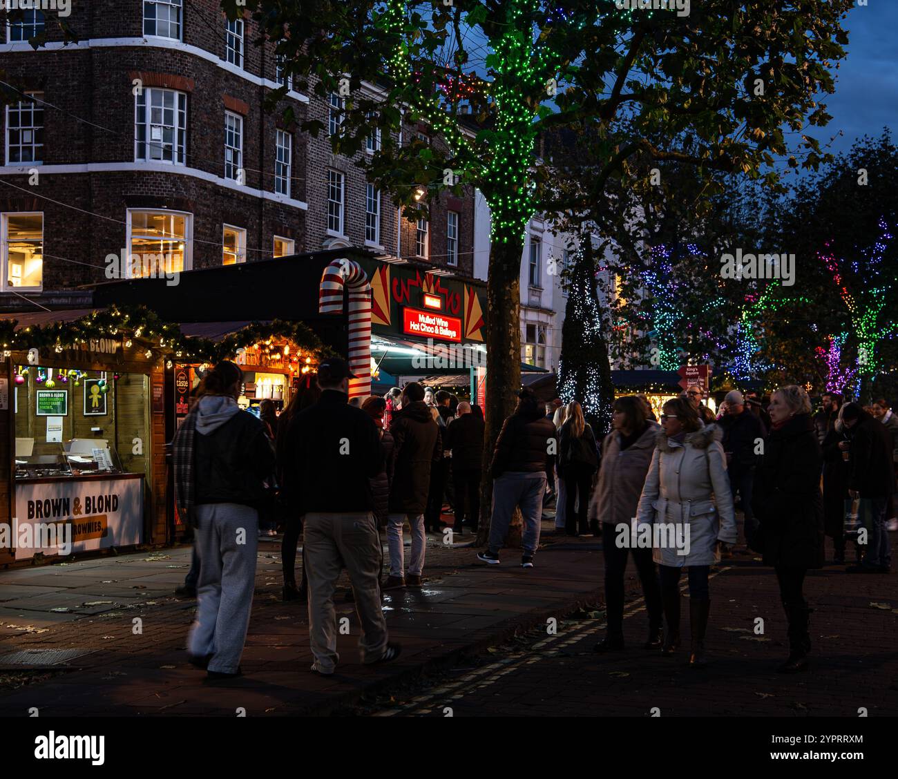 Die Menschenmassen versammeln sich am Abend auf einem Weihnachtsmarkt mit beleuchteten Bäumen und dekorierten Ständen in einer gemütlichen urbanen Umgebung. York Christmas Market 2024 Stockfoto