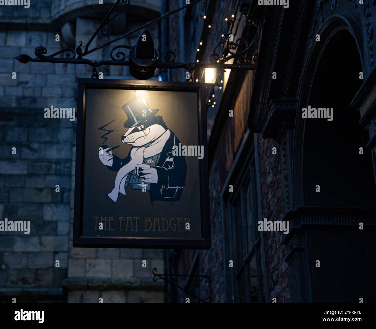 Vintage-Pub-Schild mit einem Cartoon-Dachs mit Hut und Schal, beleuchtet nachts vor einem strukturierten Backsteinbau, der den Fat Badger York trägt Stockfoto