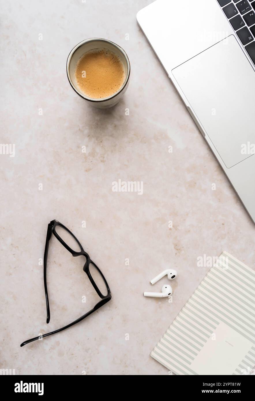 Minimaler Arbeitsplatz mit Laptop, schwarzen Gläsern, Kaffeetasse, schnurlosen Ohrstöpseln und gestreiftem Notebook auf weißem Schreibtisch – moderne professionelle Einrichtung. Draufsicht. Stockfoto