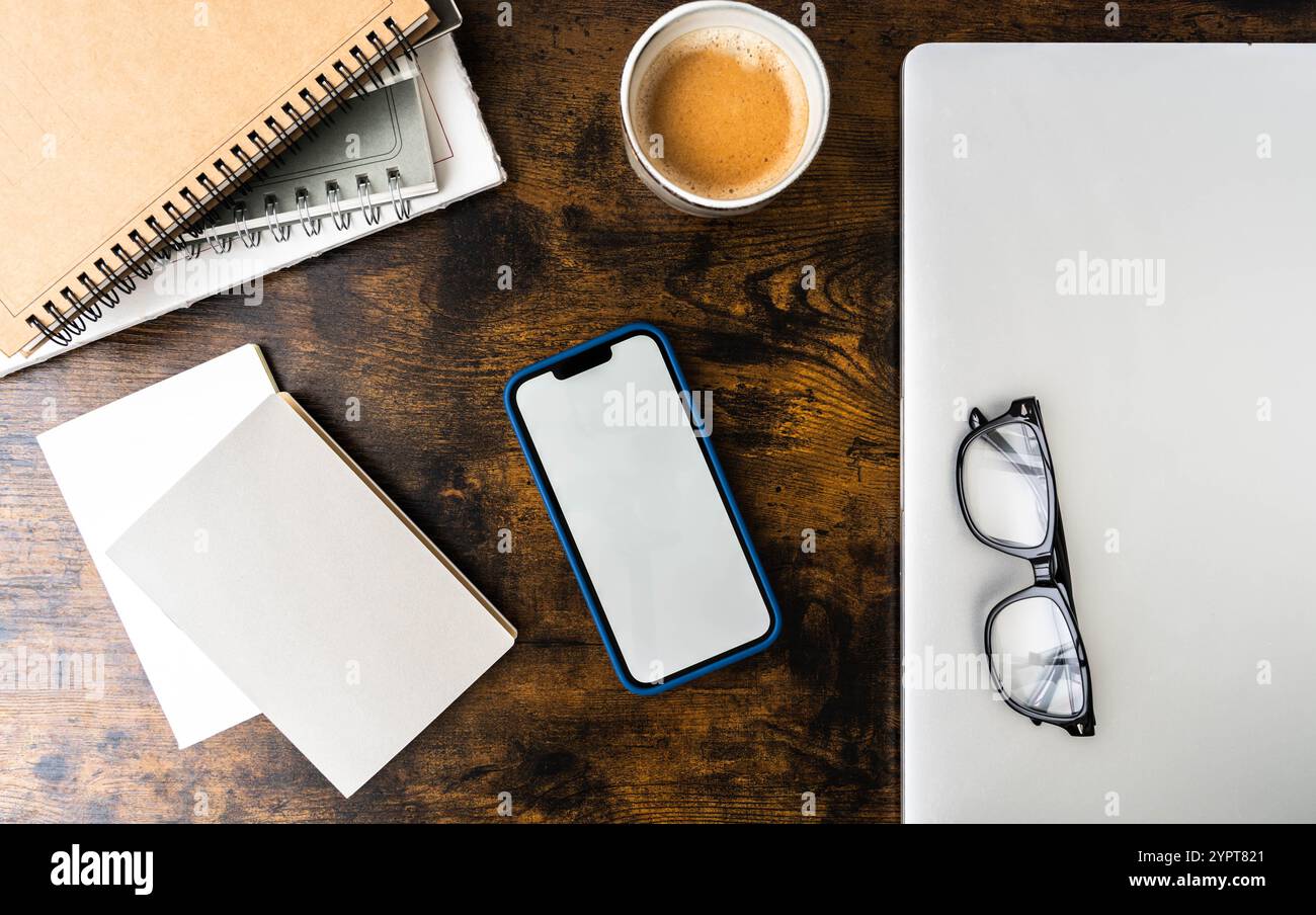 Arbeitsbereich mit Smartphone in blauer Hülle, Spiralnotizen, leerem Papier, Gläsern, Kaffeetasse, und Laptop auf Holzschreibtisch - moderne, stilvolle Einrichtung. Draufsicht Stockfoto