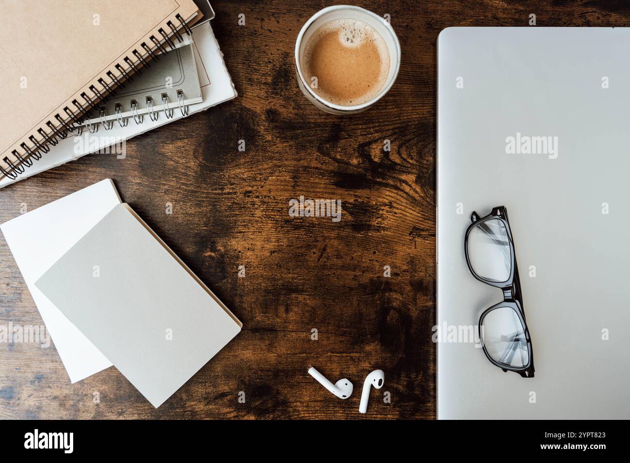 Minimaler Arbeitsplatz mit Kaffeetasse, Spiralnotizbüchern, leerem Papier, Brillen, schnurlosen Ohrstöpseln, und Holzschreibtisch - moderne, stilvolle Einrichtung. Draufsicht. Stockfoto